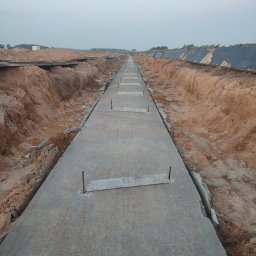 MAR-BUD Marcin Śledziński - Wąski, betonowy chodnik z metalowymi prętami, otoczony wykopami ziemnymi, ciągnący się w dal, pod pochmurnym niebem.