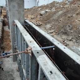 MAR-BUD Marcin Śledziński - Szare szalunki z metalowymi elementami mocującymi, przygotowane do wylania betonu. W tle widoczny wykop i usypana ziemia.  Budowa fundamentów.