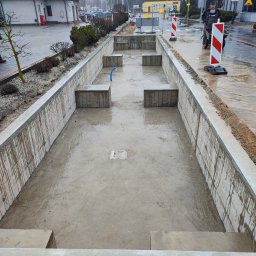 MAR-BUD Marcin Śledziński - Betonowa konstrukcja w ziemi, prawdopodobnie zbiornik retencyjny w trakcie budowy, widoczne ślady po zalaniu betonem, w tle budynki i pracownik z taczką.