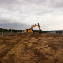 Żółta koparka JCB na placu budowy z rzędami białych słupów. Ślady opon na piasku i pochmurne niebo w tle. Etap przygotowania terenu pod budowę.