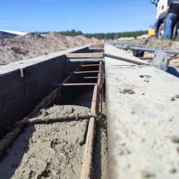 MAR-BUD Marcin Śledziński - Zalewanie fundamentów betonem w szalunku z widocznym zbrojeniem. W tle teren budowy i betoniarka. Ujęcie z niskiej perspektywy.