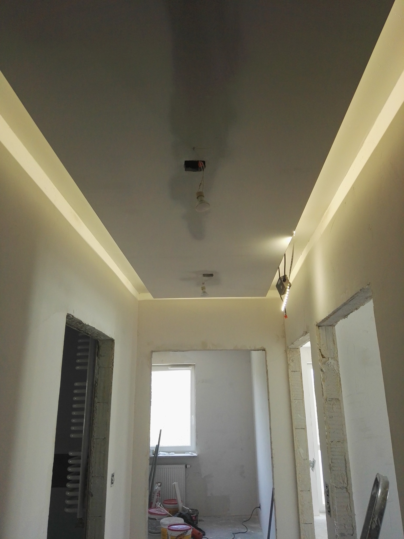 Wykończony sufit podwieszany z oświetleniem LED w korytarzu, widoczne puszki elektryczne, ściany przygotowane do malowania, surowy otwór drzwiowy.