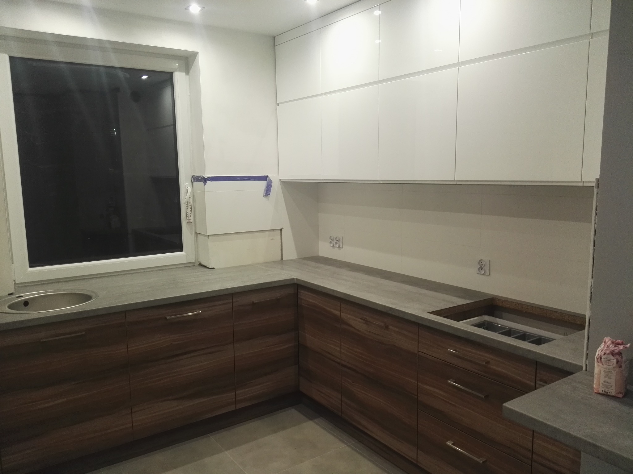 Nowoczesna kuchnia w tonacji bieli i drewna:  białe, błyszczące szafki wiszące, drewnopodobne szafki dolne, szary blat i okno. Widoczny fragment zlewu.