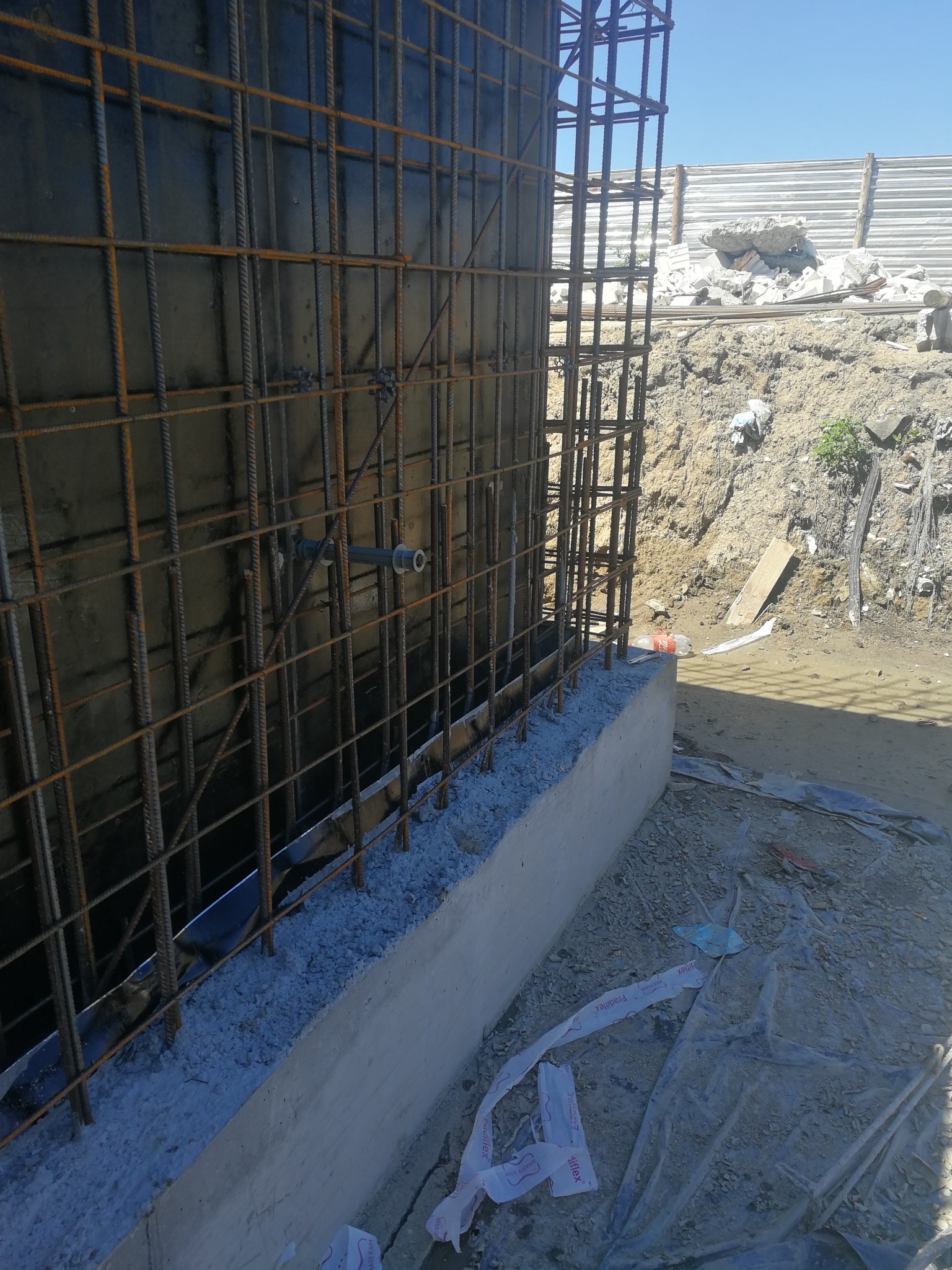 Zbrojenie pionowe ściany fundamentowej z widocznymi prętami i betonową podstawą. W tle ziemia i tymczasowe ogrodzenie placu budowy.