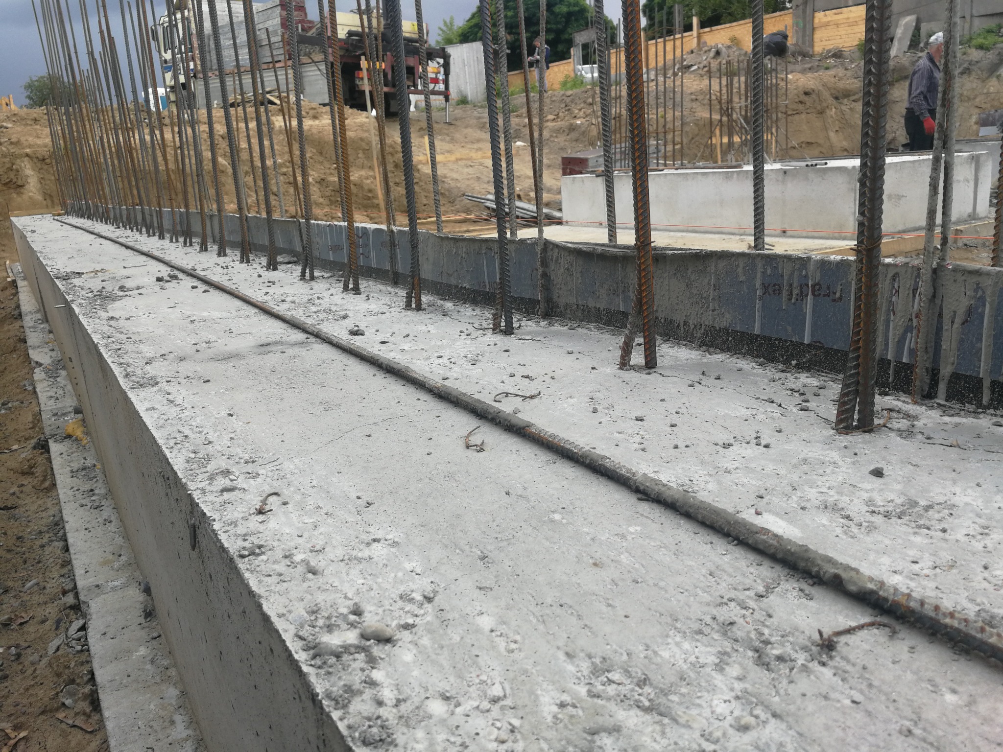 Betonowy fundament z wystającymi prętami zbrojeniowymi, widok z góry, w tle wykop i częściowo widoczny samochód ciężarowy.