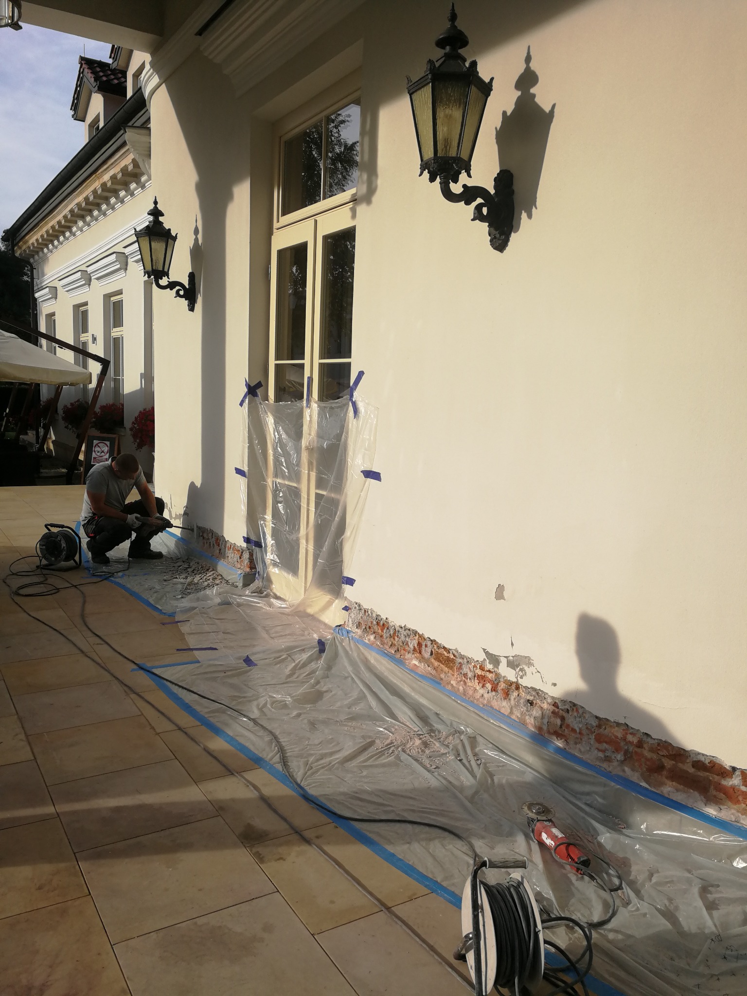 Pracownik w trakcie renowacji elewacji budynku. Oczyszczanie ściany wokół okna, zabezpieczonego folią. Widoczne narzędzia, osłony i fragmenty usuniętego tynku.