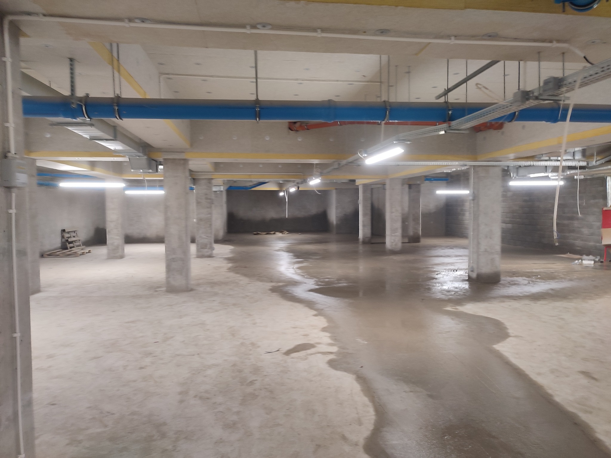 Surowy stan garażu podziemnego z widocznymi instalacjami pod stropem i betonowymi słupami. Na podłodze widoczne ślady wody.