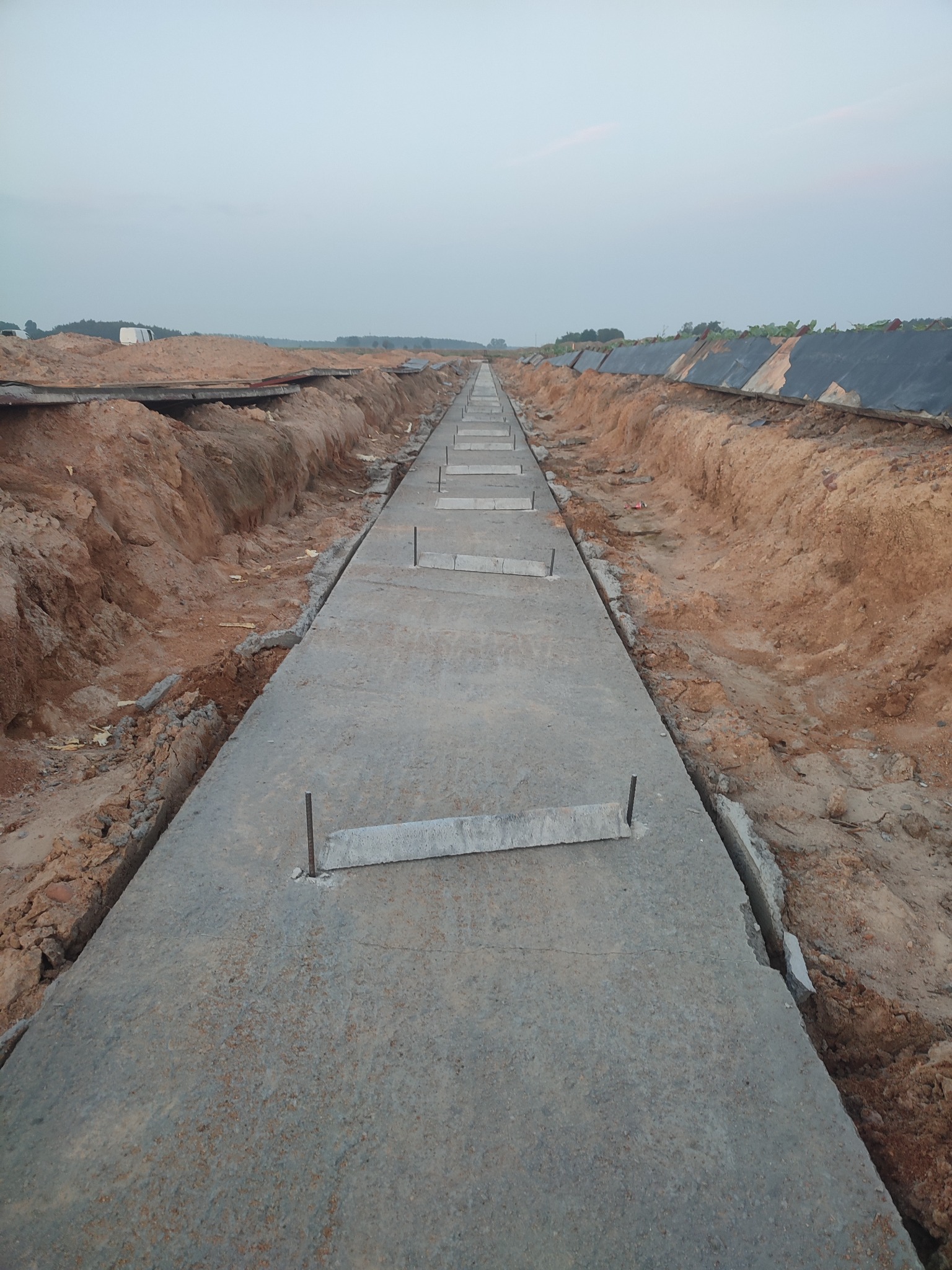 Wąski, betonowy chodnik z metalowymi prętami, otoczony wykopami ziemnymi, ciągnący się w dal, pod pochmurnym niebem.