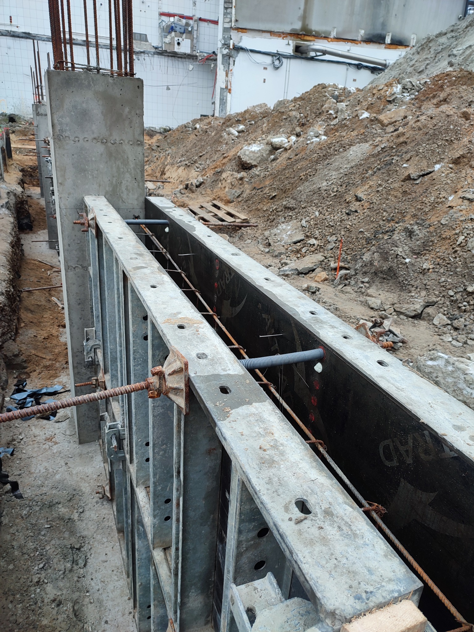 Szare szalunki z metalowymi elementami mocującymi, przygotowane do wylania betonu. W tle widoczny wykop i usypana ziemia.  Budowa fundamentów.