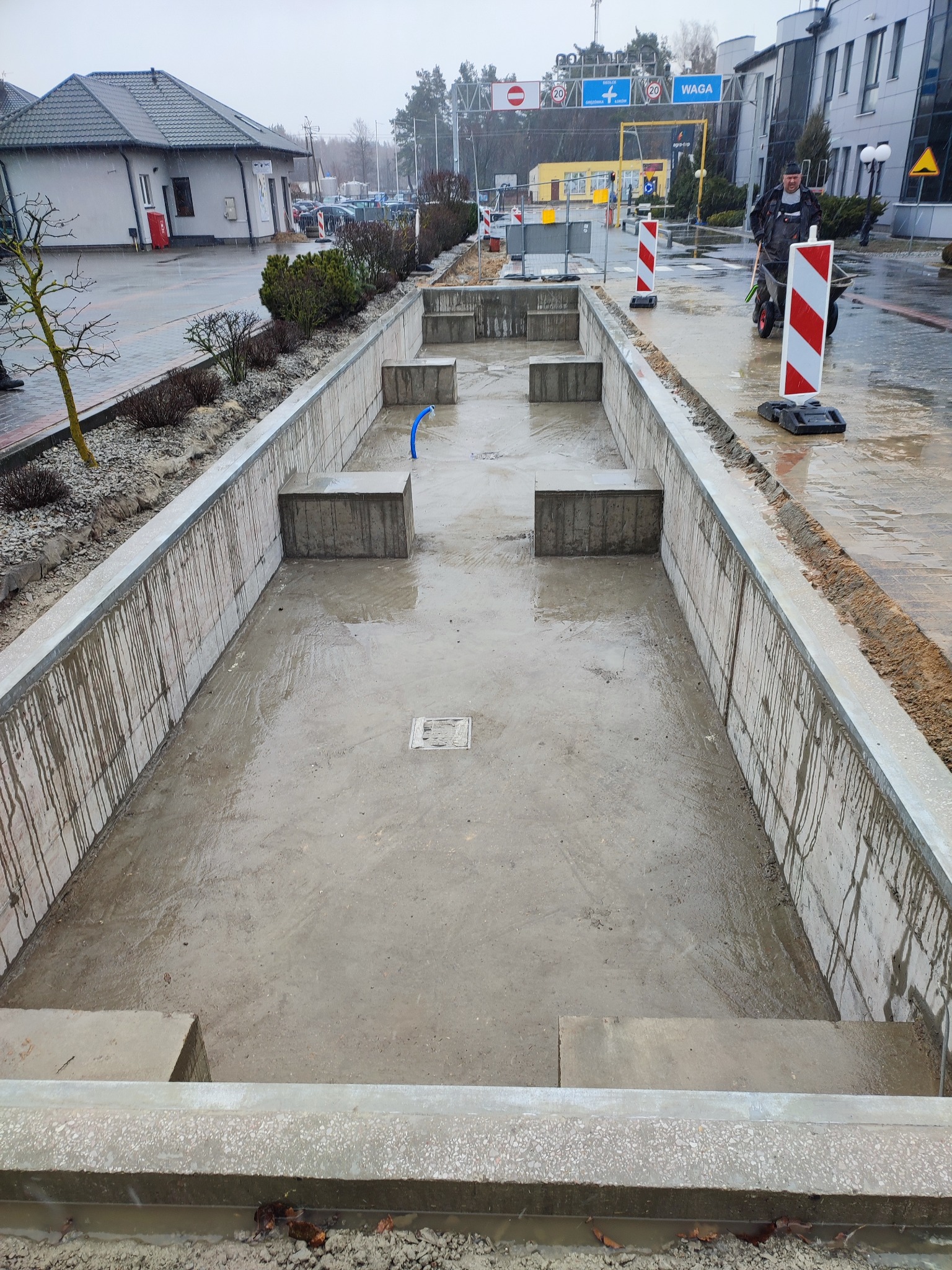 Betonowa konstrukcja w ziemi, prawdopodobnie zbiornik retencyjny w trakcie budowy, widoczne ślady po zalaniu betonem, w tle budynki i pracownik z taczką.