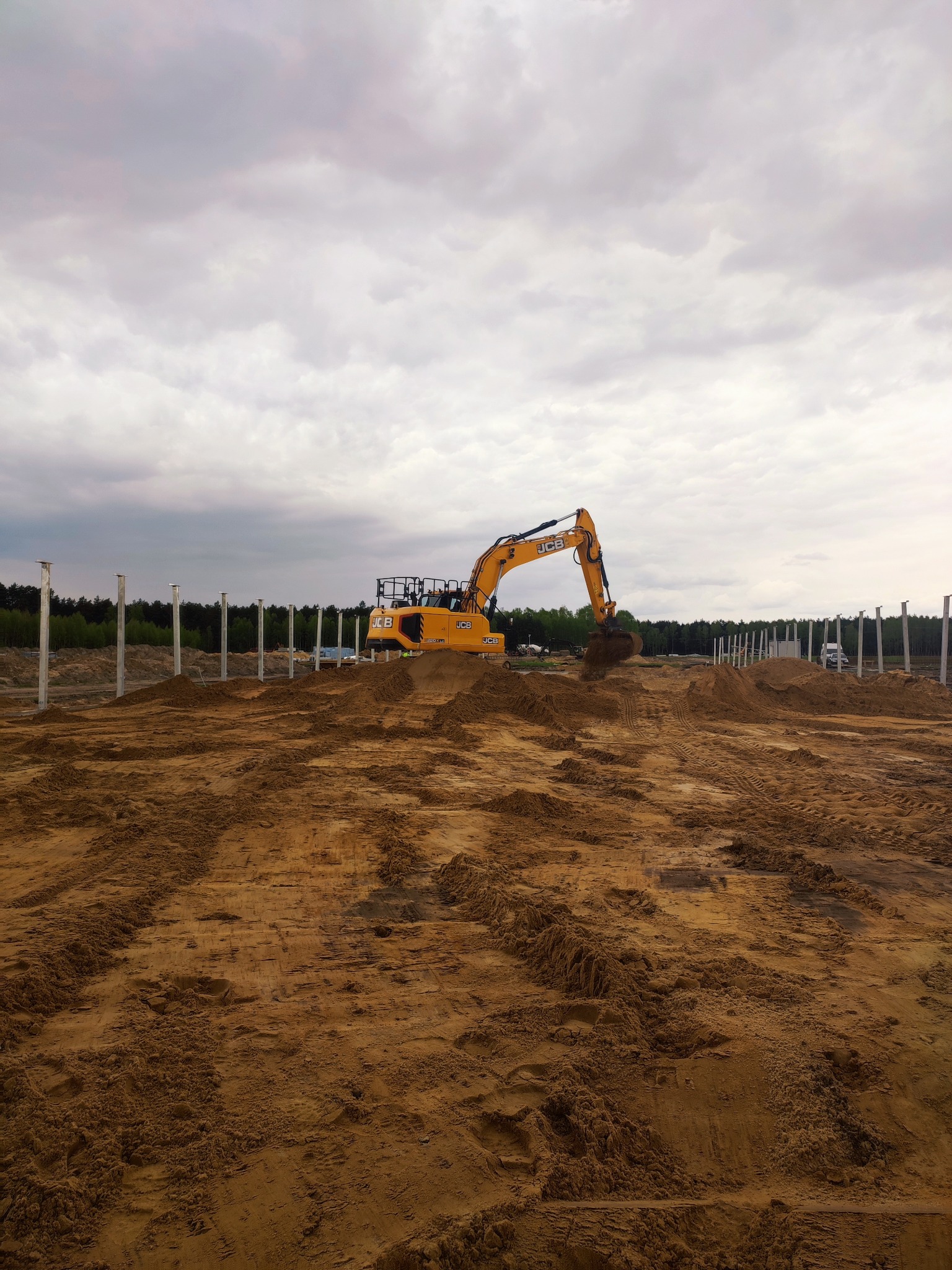 Żółta koparka JCB na placu budowy z rzędami białych słupów. Ślady opon na piasku i pochmurne niebo w tle. Etap przygotowania terenu pod budowę.