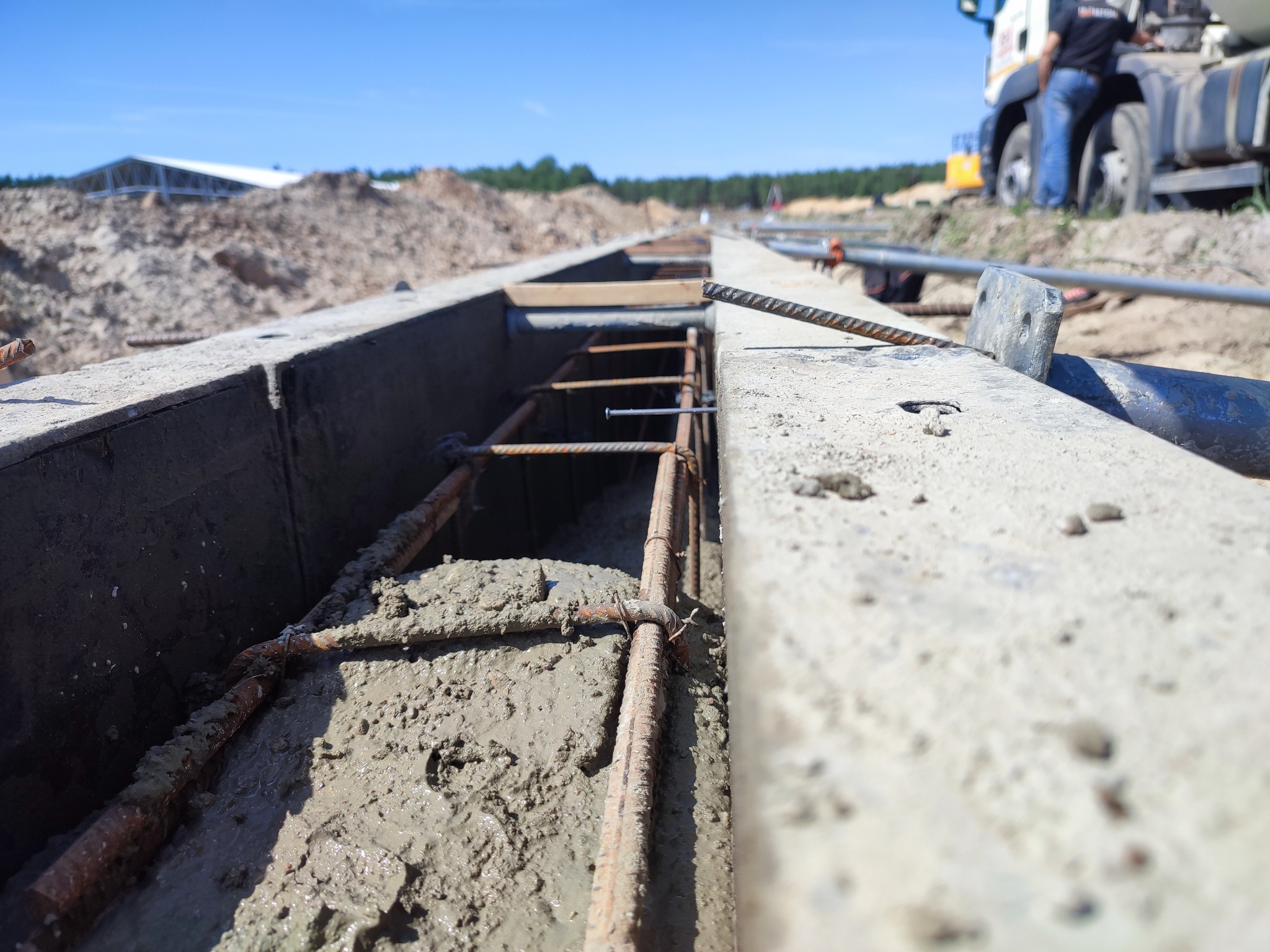 Zalewanie fundamentów betonem w szalunku z widocznym zbrojeniem. W tle teren budowy i betoniarka. Ujęcie z niskiej perspektywy.