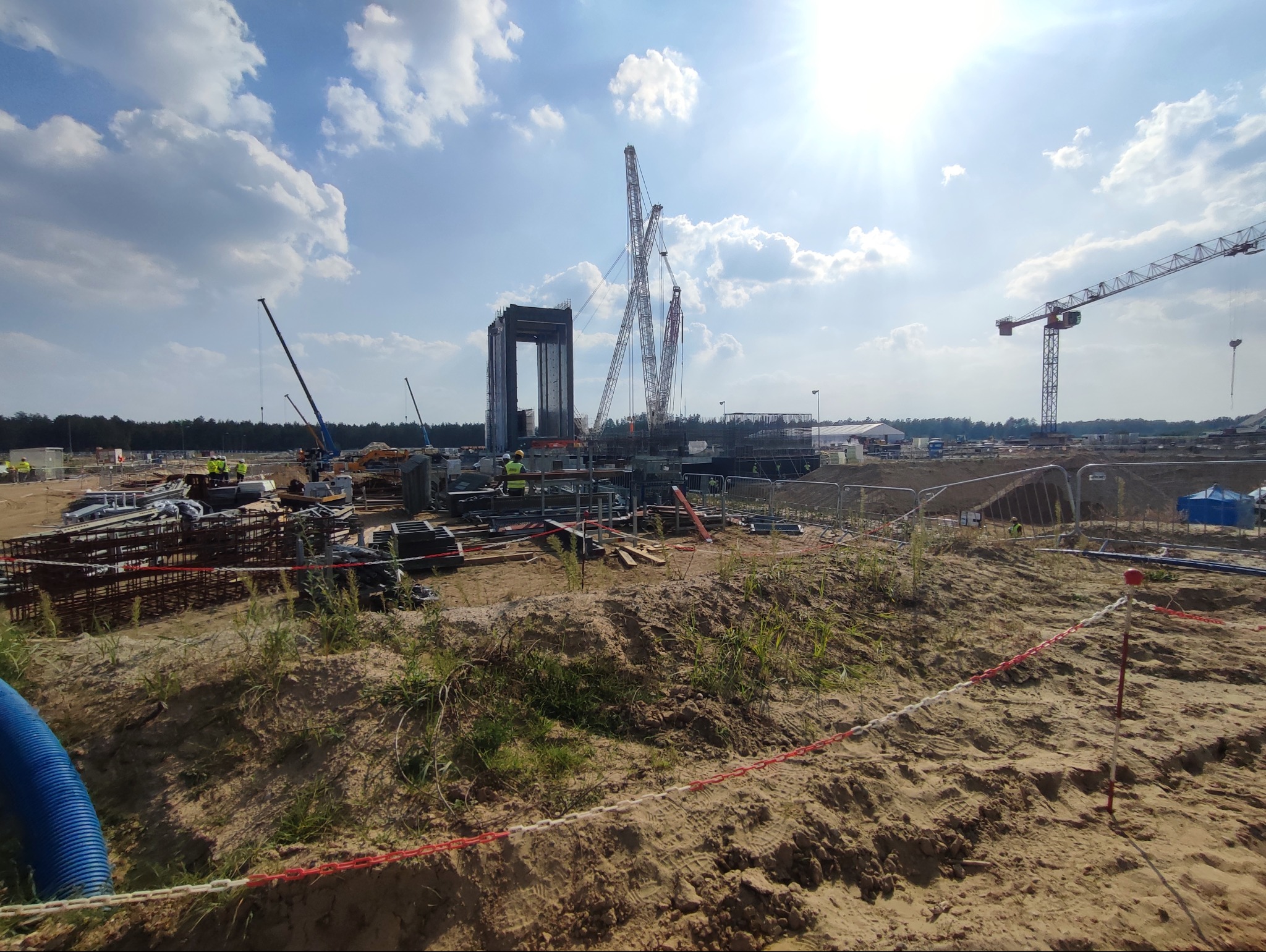 Budowa w toku: żelbetowa konstrukcja wznoszona przez zespół dźwigów na placu budowy w słoneczny dzień. Widoczne elementy zbrojenia i pracownicy w żółtych kamizelkach.