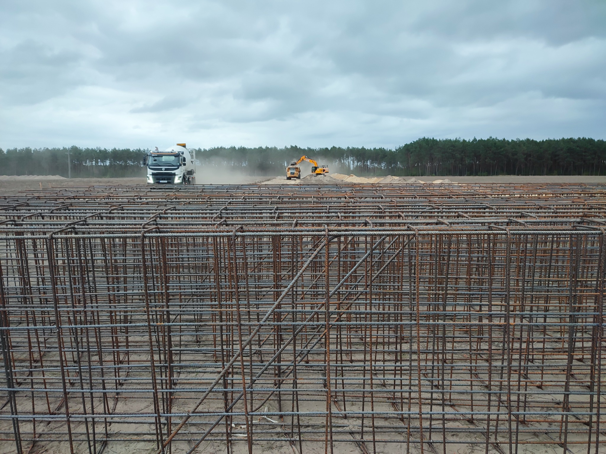 Zbrojenie fundamentów na placu budowy, w tle betoniarka i koparka. Widok na rozległy teren budowy z lasem w tle, pochmurne niebo. Zbrojenie z bliska.