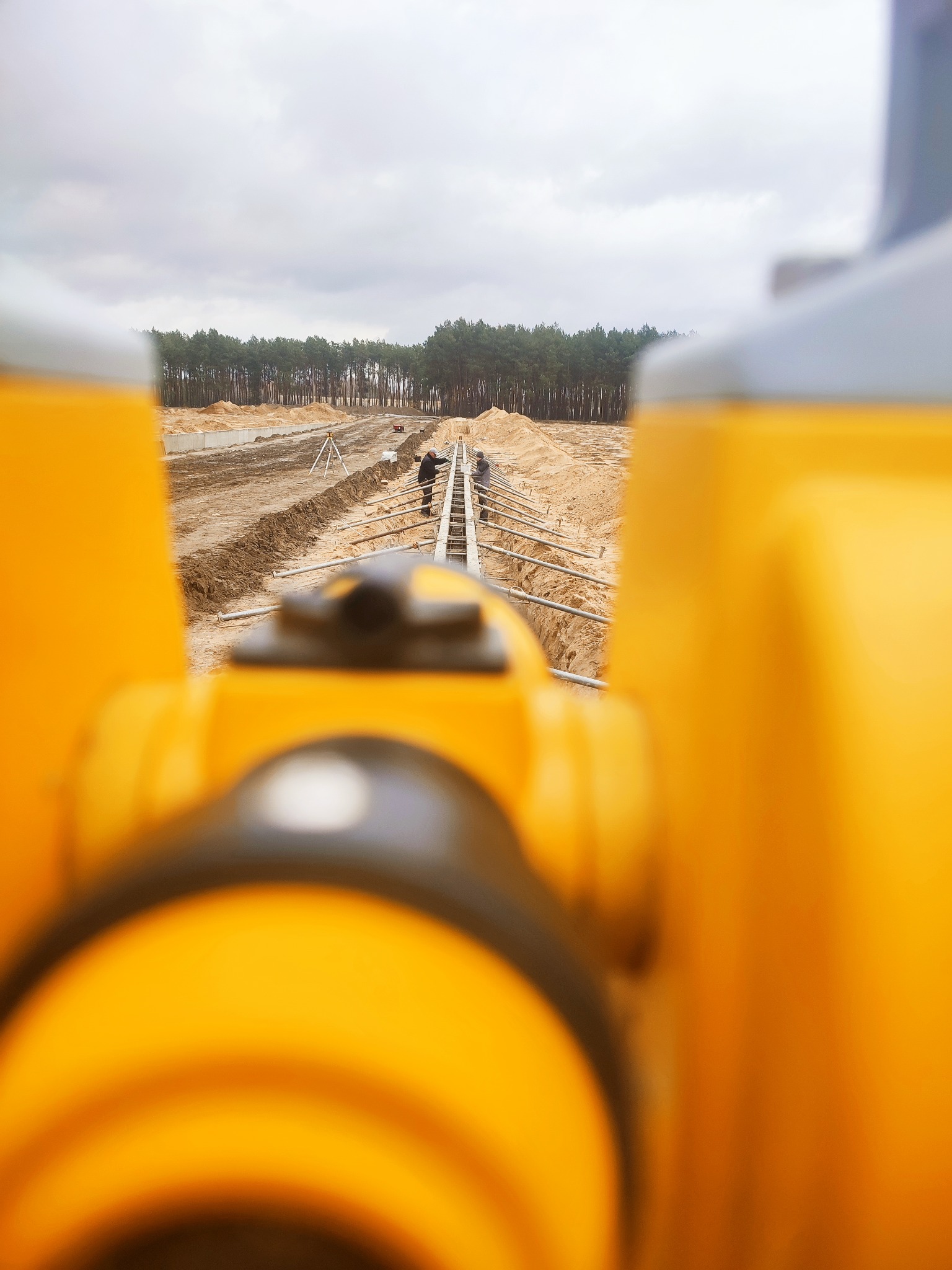 Prace ziemne pod budowę widziane przez żółty niwelator. W tle pracownicy przy wykopach i las.