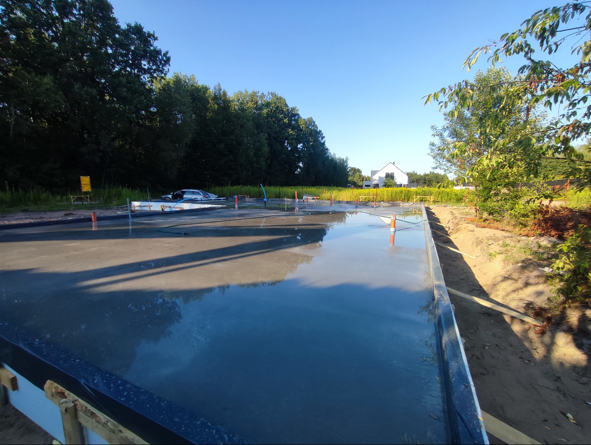 Wylany fundament pod budowę domu jednorodzinnego. Widoczne szalunki i wypoziomowany beton. W tle zieleń drzew i błękitne niebo.