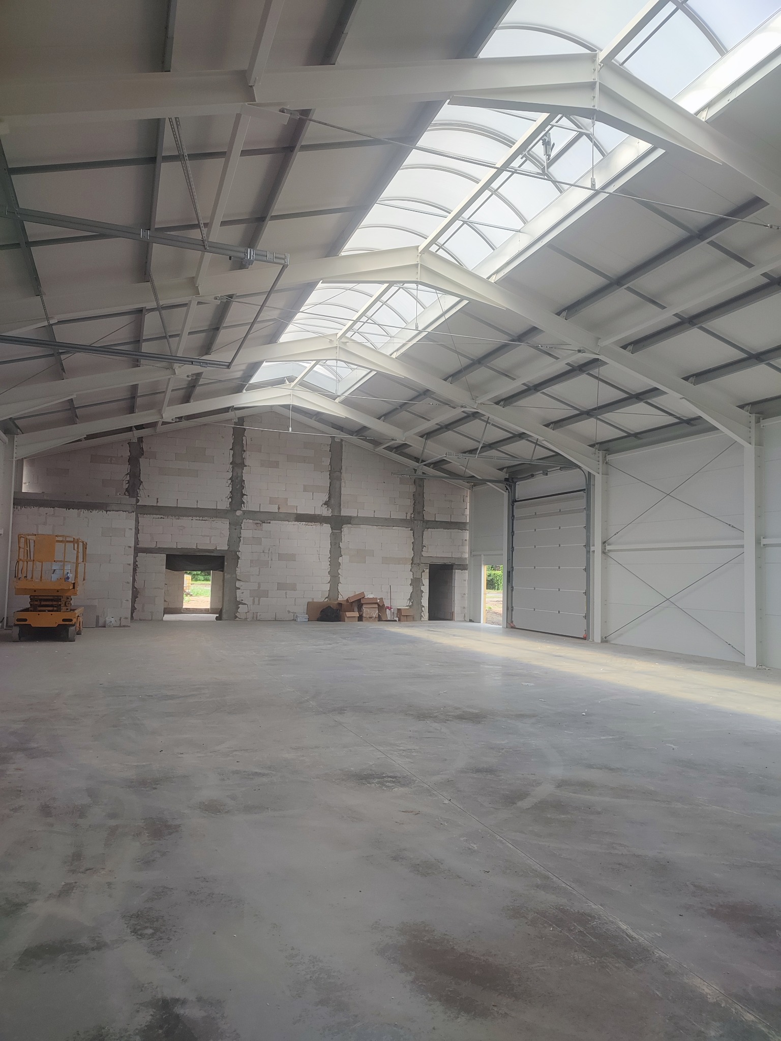 Wnętrze hali w trakcie budowy z widoczną konstrukcją stalową dachu, ścianami z bloczków i podnośnikiem koszowym. Widoczne okna dachowe i brama wjazdowa.
