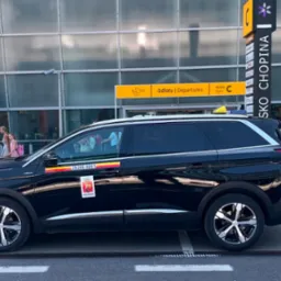 Czarny minivan taxi z logo Warszawy na tle lotniska Chopina, z pasażerami i bagażami, w słoneczny dzień.