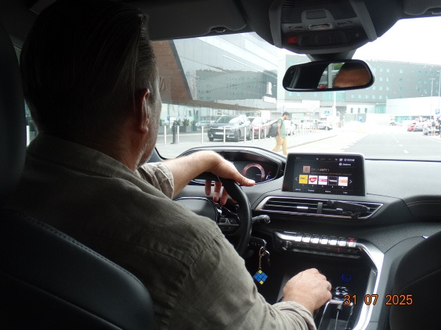 Wnętrze samochodu osobowego z kierowcą za kierownicą, widok z tylnego siedzenia, z widocznym budynkiem lotniska w tle. Na desce rozdzielczej widoczny wyświetlacz.