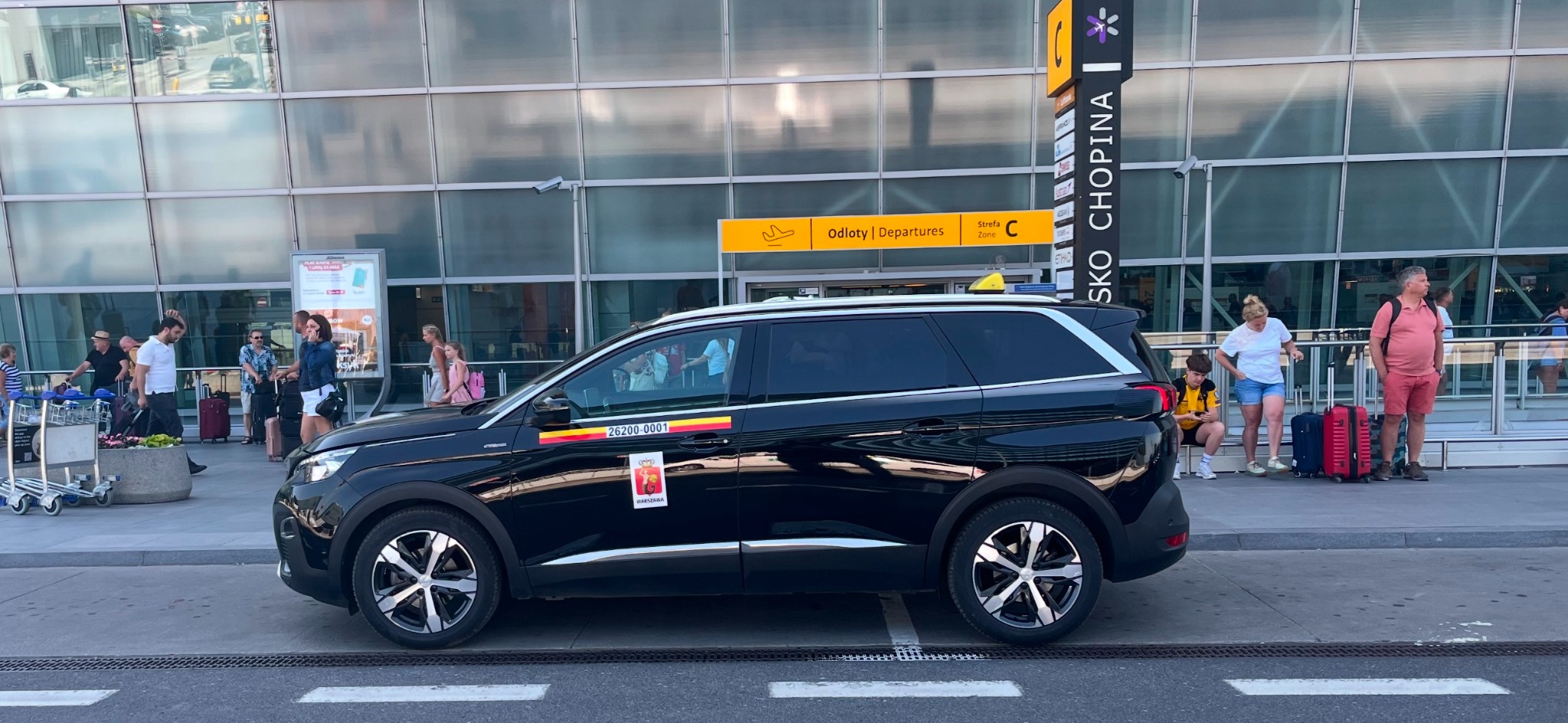 Czarny minivan taxi z logo Warszawy na tle lotniska Chopina, z pasażerami i bagażami, w słoneczny dzień.