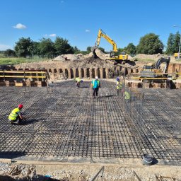 SPEC-GROUP SPÓŁKA Z OGRANICZONĄ ODPOWIEDZIALNOŚCIĄ - Rozległe zbrojenie fundamentów pod budowę, z pracownikami w kamizelkach i kaskach, oraz koparkami w tle. Widok na plac budowy w słoneczny dzień.