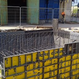 SPEC-GROUP SPÓŁKA Z OGRANICZONĄ ODPOWIEDZIALNOŚCIĄ - Zbrojenie i szalunek fundamentów z żółtymi panelami Doka, widoczny pracownik w kasku. Stalowe pręty zbrojeniowe tworzą siatkę wewnątrz szalunku, przygotowane do zalania betonem.