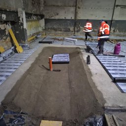 SPEC-GROUP SPÓŁKA Z OGRANICZONĄ ODPOWIEDZIALNOŚCIĄ - Przygotowanie podłoża pod fundamenty: wykop z rurą, izolacja termiczna 'Fundament 4.0 Szybki Profil SBS', robotnicy w odblaskowych ubraniach. Wnętrze budynku w trakcie prac.