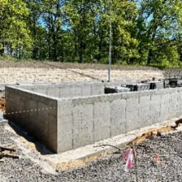 Betonowe ściany fundamentowe w wykopie, widoczne narzędzia i poziomica. Tło: drzewa i nasyp. Ujęcie z bliska, oświetlenie dzienne.