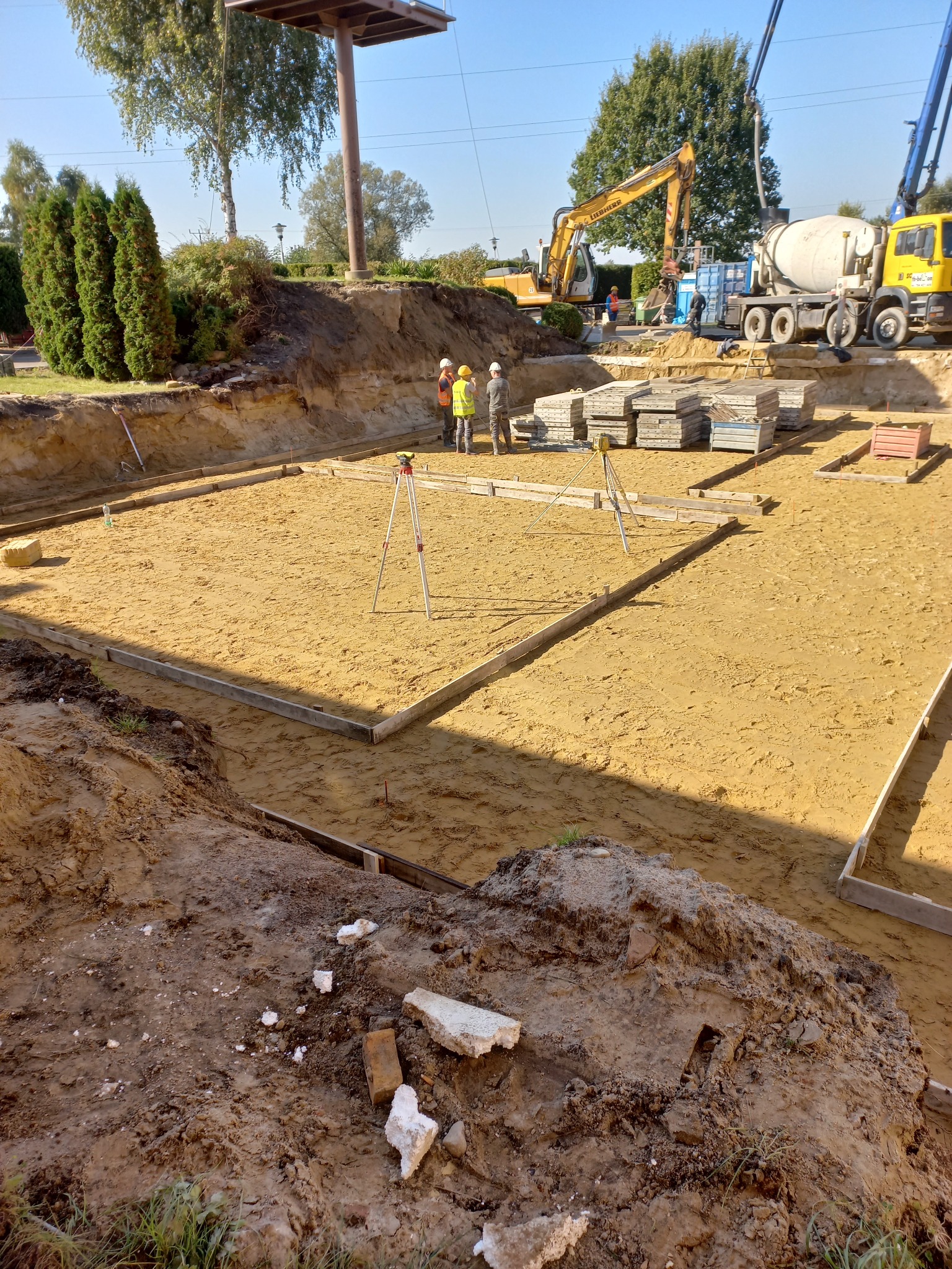 Przygotowany wykop pod fundamenty z drewnianym szalunkiem, niwelatorami i pracownikami w kaskach. W tle ciężki sprzęt budowlany: koparka i betoniarka.