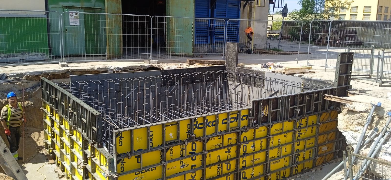 Zbrojenie i szalunek fundamentów z żółtymi panelami Doka, widoczny pracownik w kasku. Stalowe pręty zbrojeniowe tworzą siatkę wewnątrz szalunku, przygotowane do zalania betonem.