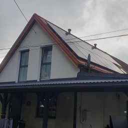 Norbud - Dom z jasną elewacją i spadzistym dachem pokrytym czerwoną blachodachówką, z zainstalowanymi panelami fotowoltaicznymi. Poniżej ciemna wiata z drewnianymi belkami i czarnymi rynnami.