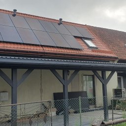 Norbud - Czerwony dach domu w Pisz z nowoczesnymi panelami fotowoltaicznymi, oknem dachowym i anteną satelitarną. Pod nim ciemna drewniana wiata z falistym zadaszeniem.