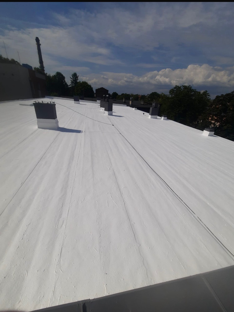 Biała, płaska powierzchnia dachu z kominami wentylacyjnymi na tle błękitnego nieba z chmurami. Widok z góry, podkreślający gładką strukturę pokrycia.