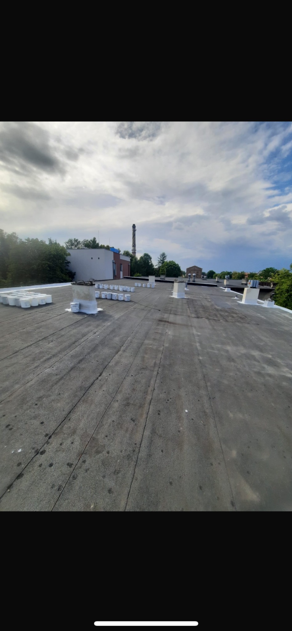 Bitumiczny dach z kominami wentylacyjnymi po malowaniu, z widocznymi puszkami farby. W tle budynek przemysłowy i błękitne niebo z chmurami.