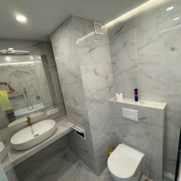 Tomasz Wojnarowicz - Wykończona łazienka w szaro-białej kolorystyce z umywalką nablatową, toaletą i lustrem, w którym odbija się osoba robiąca zdjęcie. Minimalistyczny design, płytki imitujące marmur.