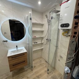 Tomasz Wojnarowicz - Nowoczesna łazienka po remoncie: kabina prysznicowa ze szklanymi drzwiami, umywalka z szafką i okrągłe lustro z podświetleniem LED. Widoczny bojler i półki wnękowe.
