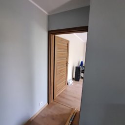 Tomasz Wojnarowicz - Świeżo pomalowane na szaro ściany w pokoju z otwartymi drewnianymi drzwiami, widoczne narzędzia malarskie i podłoga. Prace wykończeniowe w trakcie realizacji.