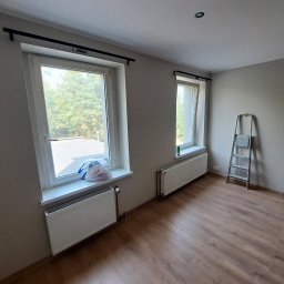 Tomasz Wojnarowicz - Jasne wnętrze po malowaniu ścian na szaro, z drewnianą podłogą i grzejnikami pod oknami. Drabina oparta o ścianę.