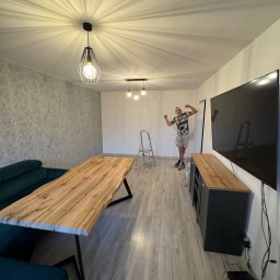 Tomasz Wojnarowicz - Jasne wnętrze po remoncie: drewniany stół, sofa, szafka RTV, telewizor. Na ścianie wzorzysta tapeta. Mężczyzna pozuje na tle drabiny.