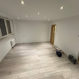 Tomasz Wojnarowicz - Jasne panele podłogowe ułożone w pustym pokoju z białymi ścianami i oknem. Widoczne narzędzia i skrzynka narzędziowa, sugerujące etap wykończenia wnętrza.