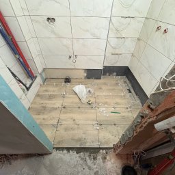 Tomasz Wojnarowicz - Układanie płytek podłogowych imitujących drewno w łazience, z krzyżykami dystansowymi. Widoczne płytki ścienne, poziomice i narzędzia glazurnicze. Prace wykończeniowe w toku.