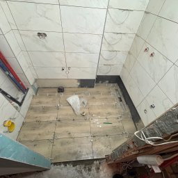 Tomasz Wojnarowicz - Wnętrze łazienki w trakcie prac glazurniczych: białe płytki marmurowe na ścianach, drewnopodobne płytki na podłodze, poziomice, krzyżyki dystansowe, widoczne instalacje.