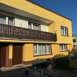 Tomasz Wojnarowicz - Żółta elewacja dwupiętrowego domu z balkonem, brązowe okna i drzwi. Widoczne ślady farby i narzędzia malarskie, sugerujące świeżo zakończone malowanie.