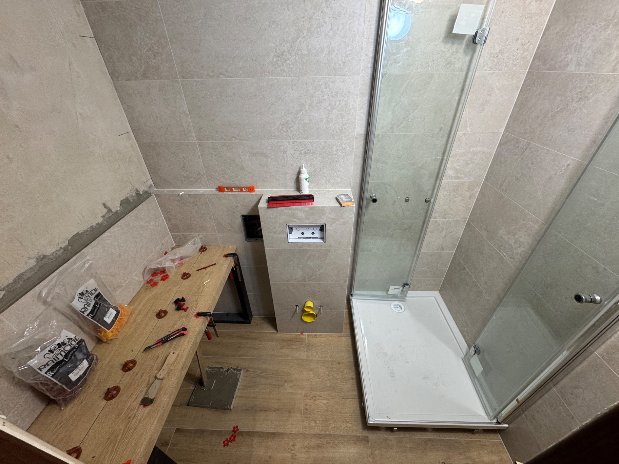 Remont łazienki w trakcie: ułożone płytki ścienne i podłogowe, zamontowany stelaż WC, kabina prysznicowa. Widoczne narzędzia glazurnicze i poziomica.