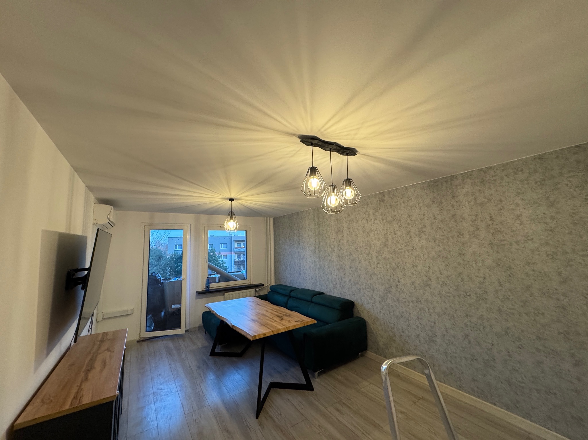 Jasne wnętrze salonu z drewnianym stołem, zieloną sofą i designerskimi lampami. Ściana pokryta tapetą o subtelnej fakturze. Widoczna drabina malarska.