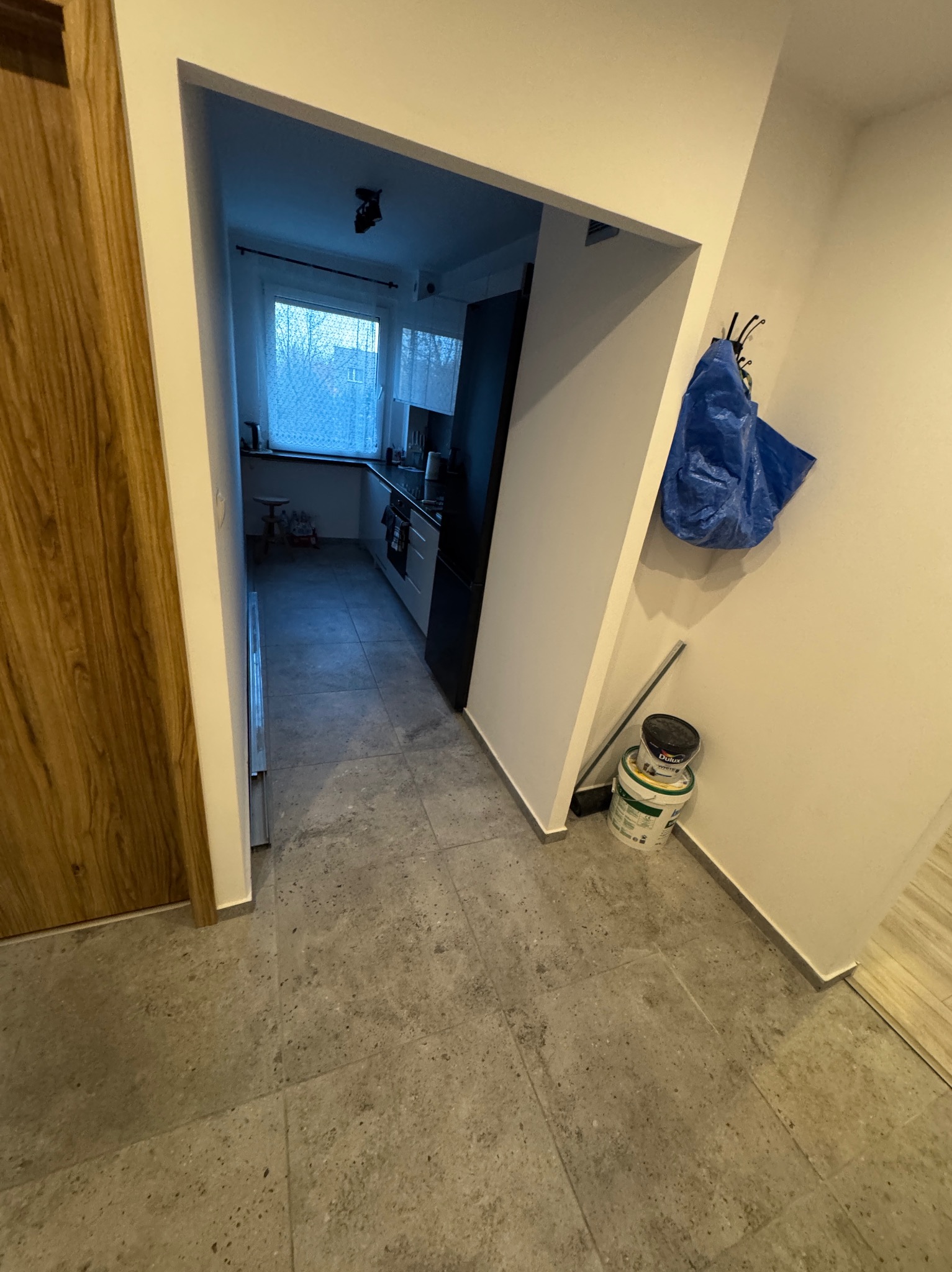Wykończone wnętrze mieszkania z widokiem na kuchnię, szare płytki podłogowe, białe ściany, drzwi z drewnianą okleiną. Widoczne wiaderka z farbą i niebieska torba.