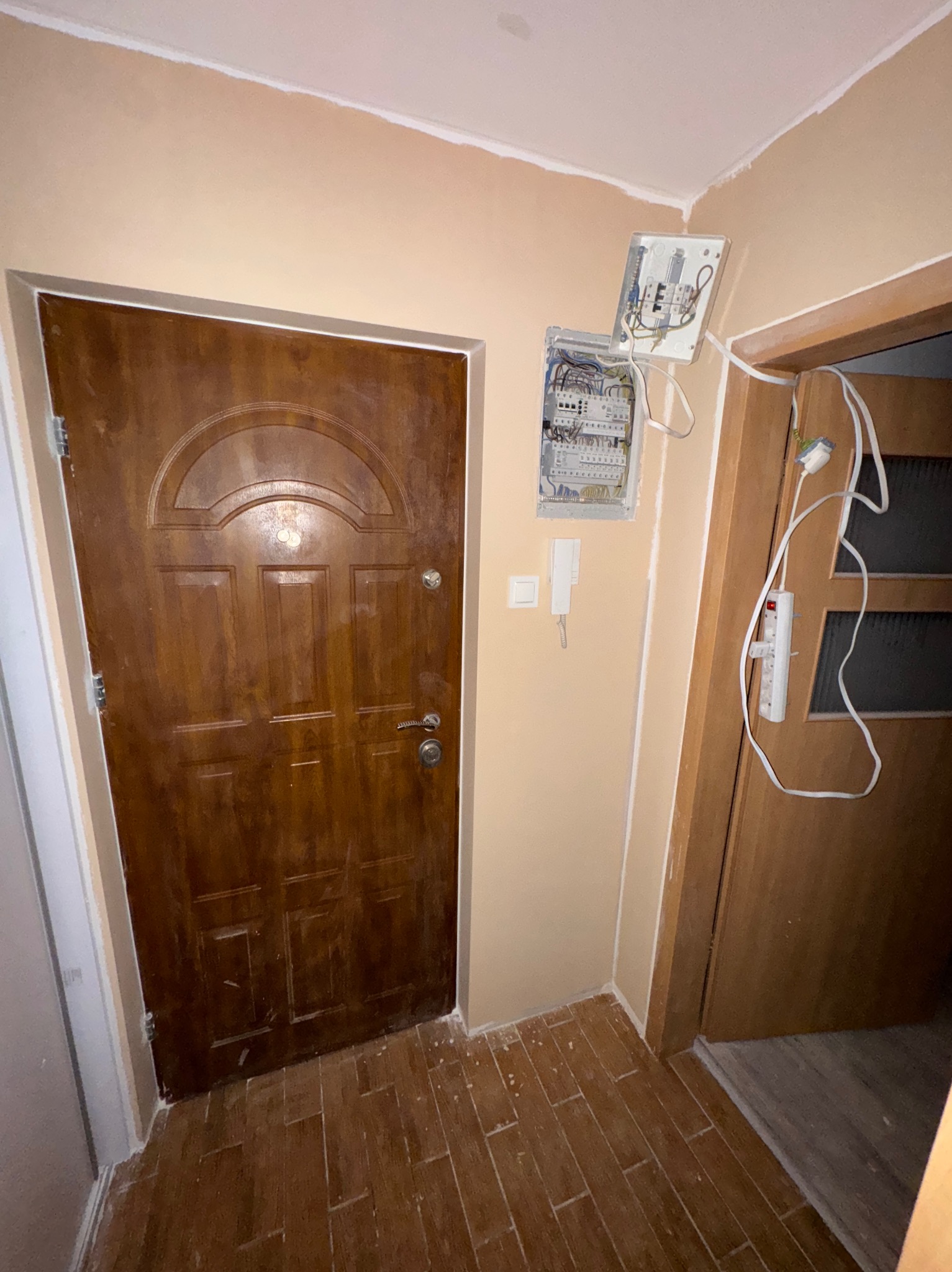 Korytarz w trakcie remontu: widoczna otwarta skrzynka elektryczna, pomalowane ściany na beżowo, drewniane drzwi wejściowe i częściowo ułożona podłoga z płytek imitujących drewno.