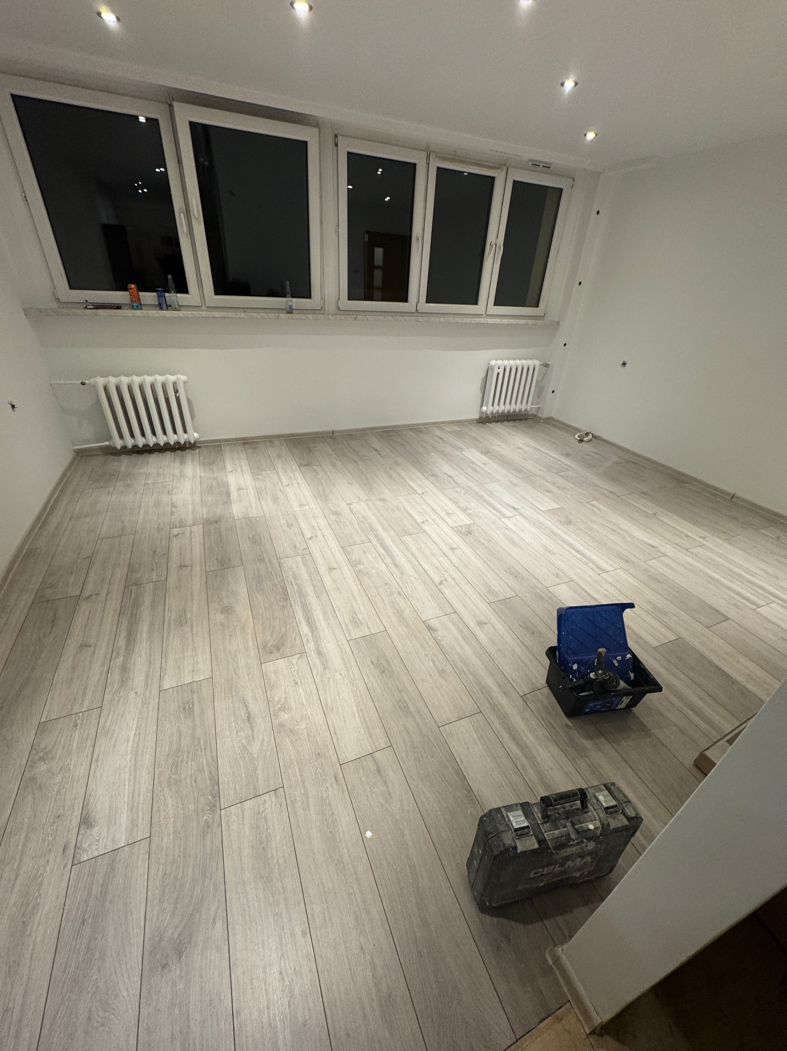 Jasne panele podłogowe ułożone w pokoju z białymi ścianami i grzejnikami. Dwa okna w tle. Na podłodze skrzynki z narzędziami.