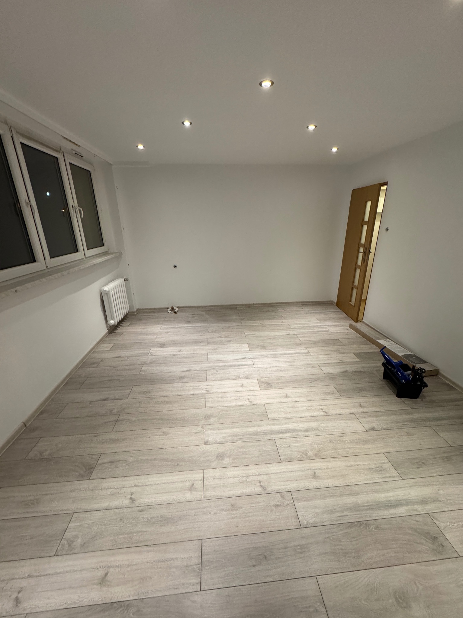 Jasne panele podłogowe w nowym, pustym pokoju z białymi ścianami i oknem. Widoczny grzejnik i narzędzie do cięcia paneli przy drzwiach.