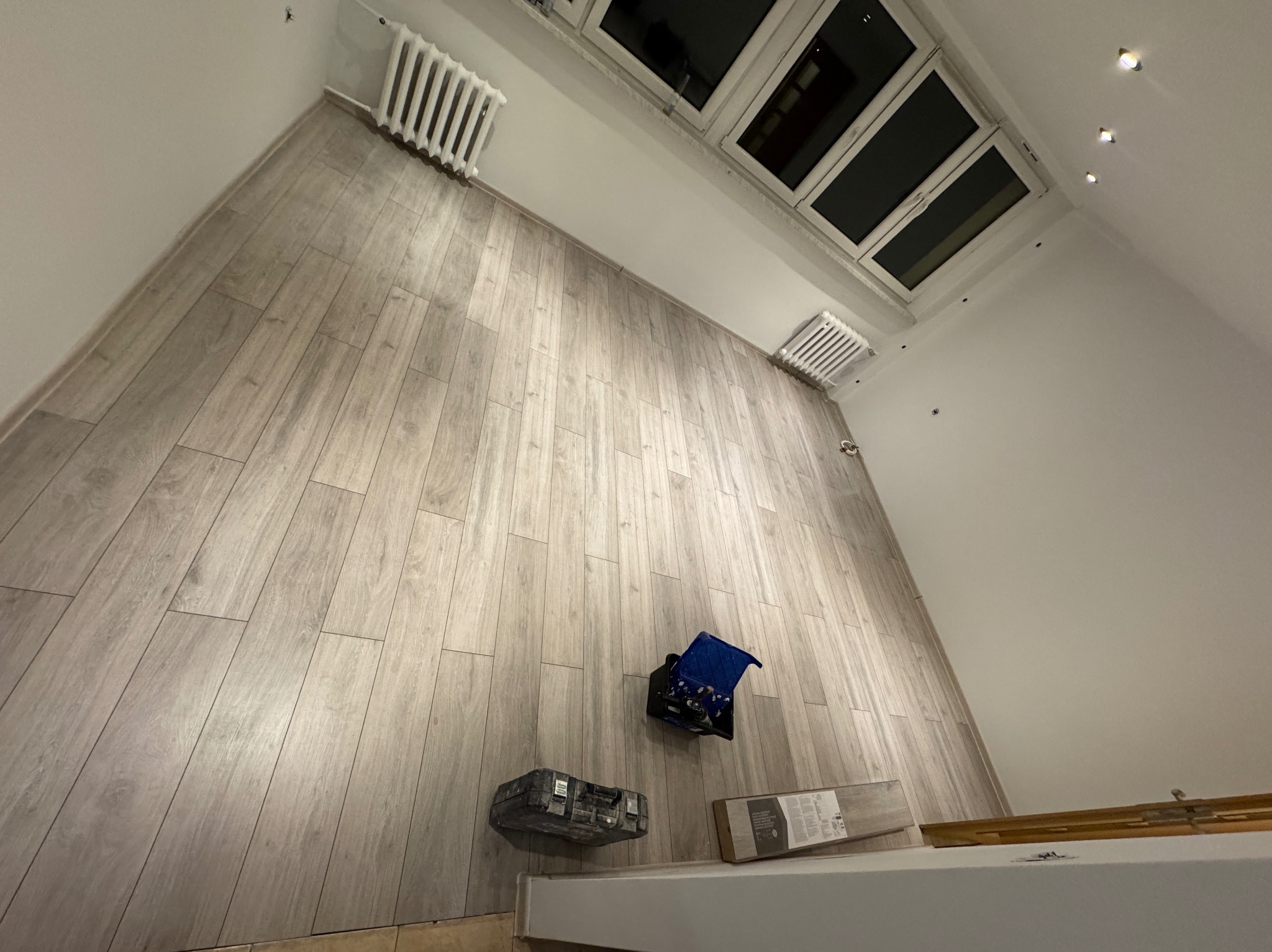 Ukończona podłoga z paneli w jasnym odcieniu drewna, widok z góry, narzędzia glazurnicze w rogu. Grzejniki pod oknami dachowymi.